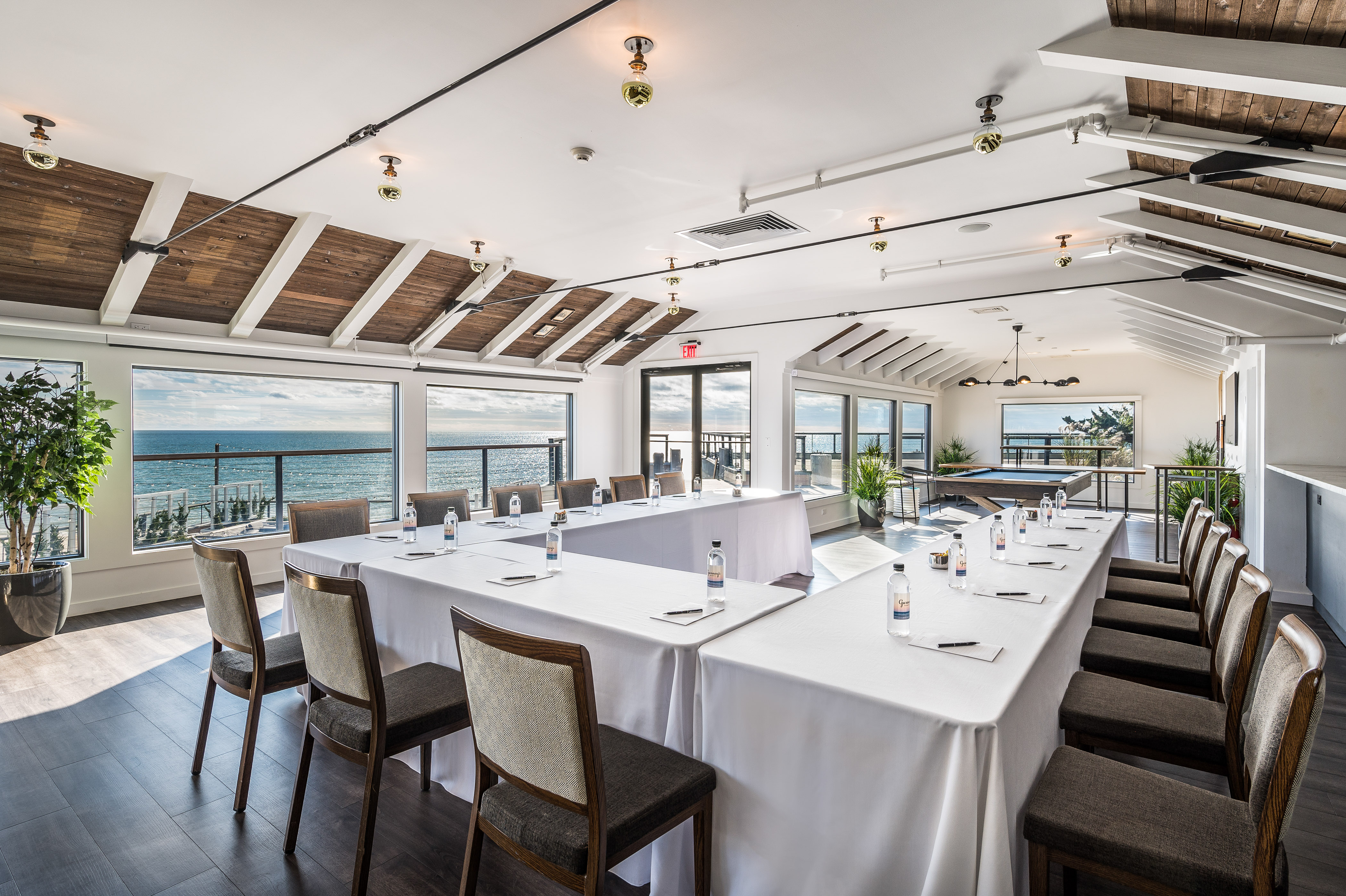 Regent Lounge with wood flooring, white walls and ceiling with wood panel accents, windows with ocean view and u-shaped table setup with white linen and wooden chairs.