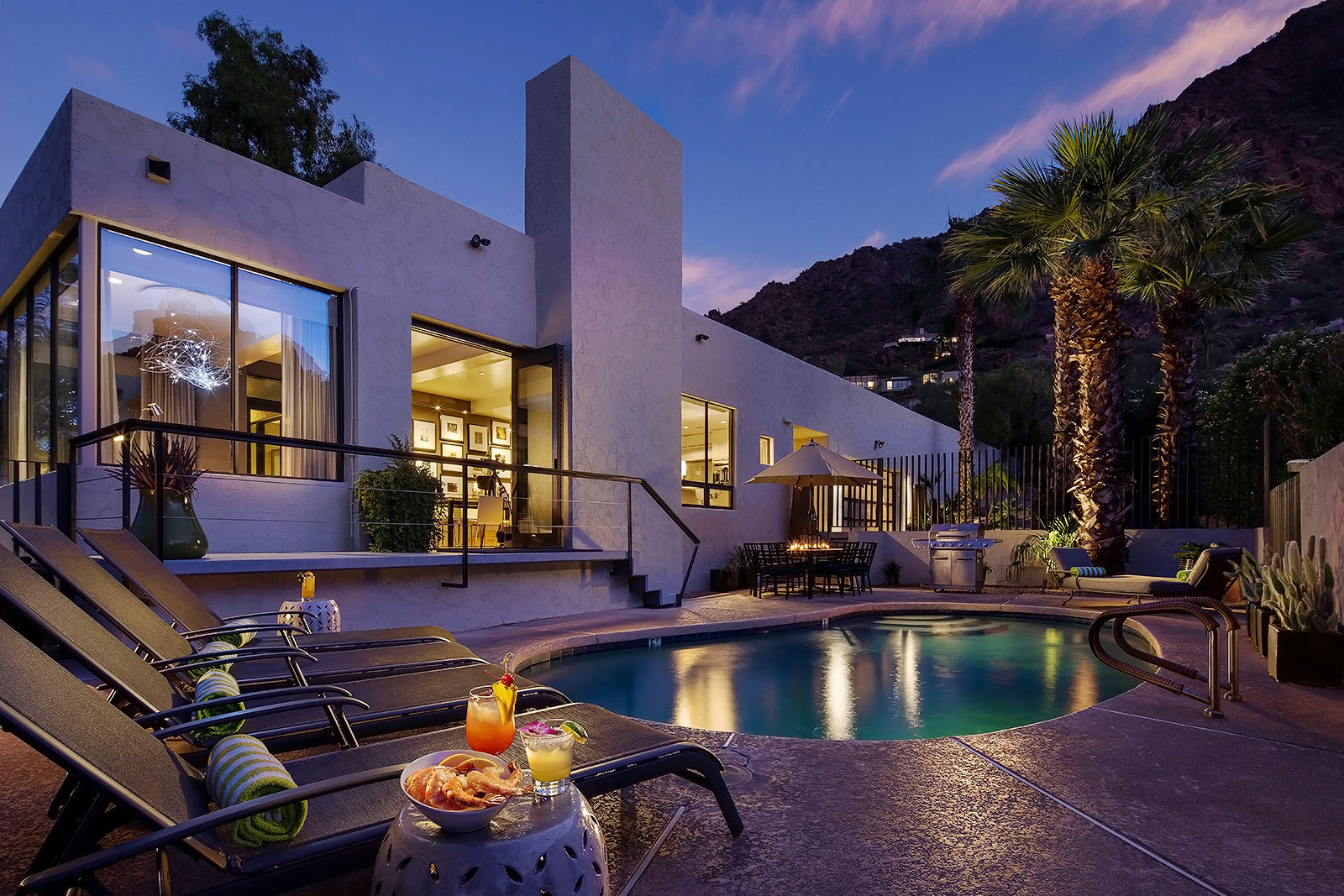 View of pool with lounge chairs, shrimp cocktail and drink at dusk, with mountainside in background.