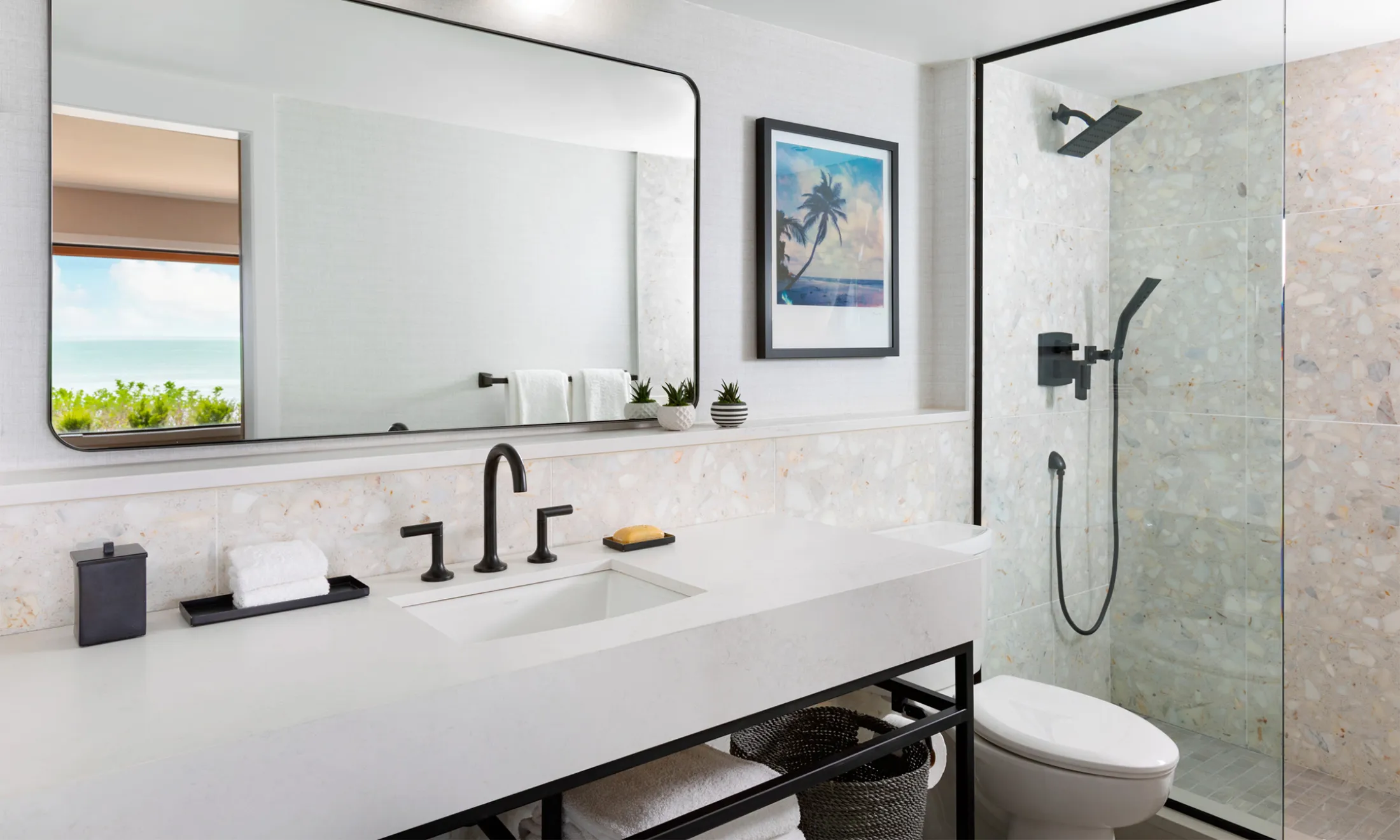 A modern bathroom with a large mirror, white sink, and glass-enclosed shower, featuring a black faucet and fixtures, with a view of the ocean in the mirror.