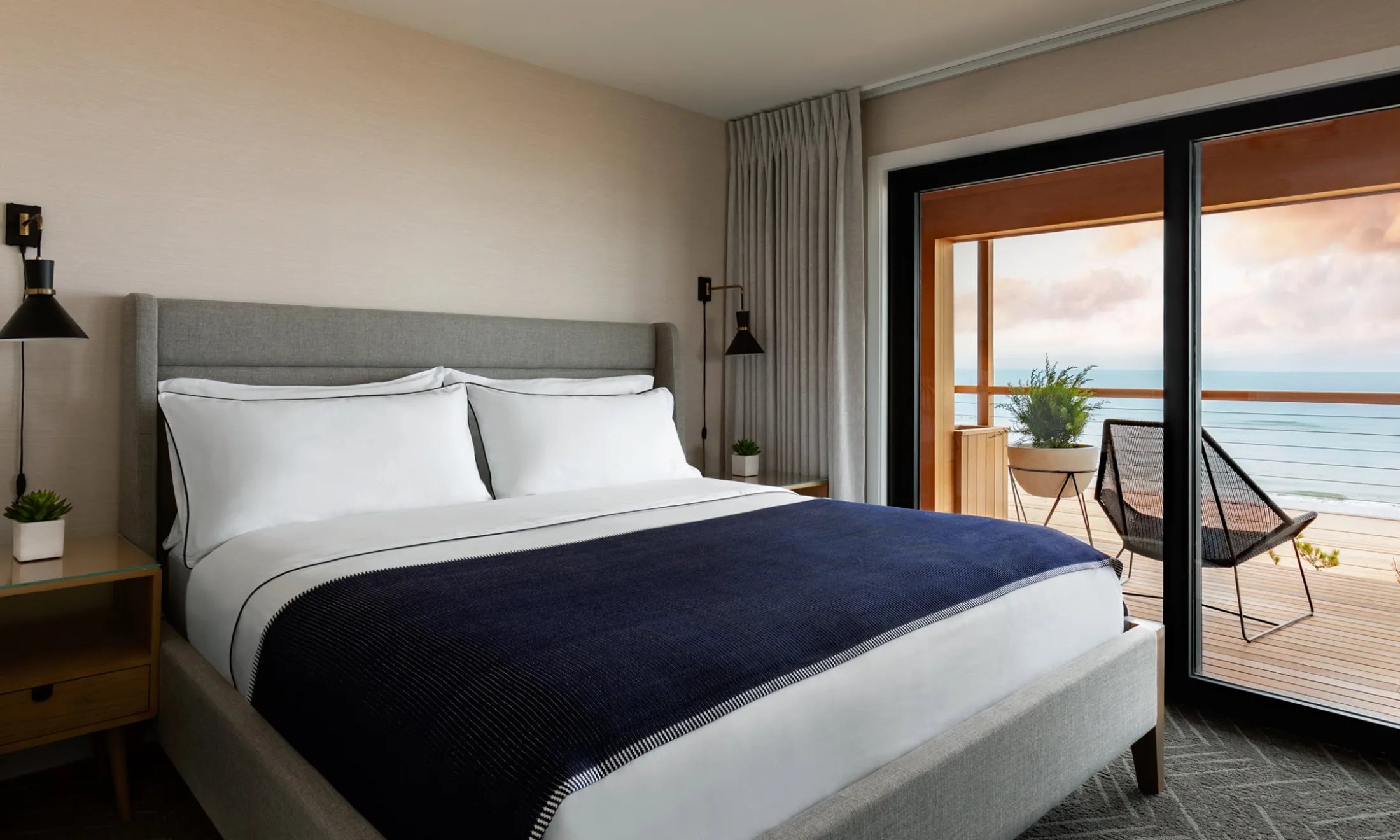A wide view of a hotel room with a bed, nightstand, lamp, chair, and sliding glass door leading to a balcony with an ocean view.