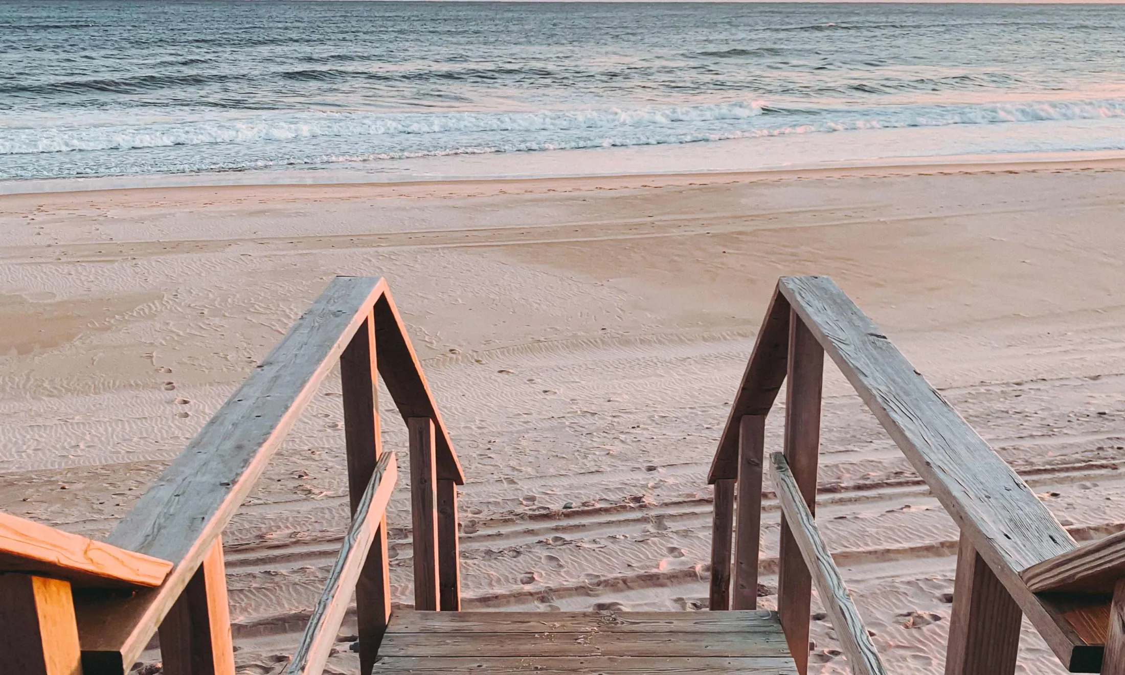 A wooden staircase leads to a beach with waves in the background.