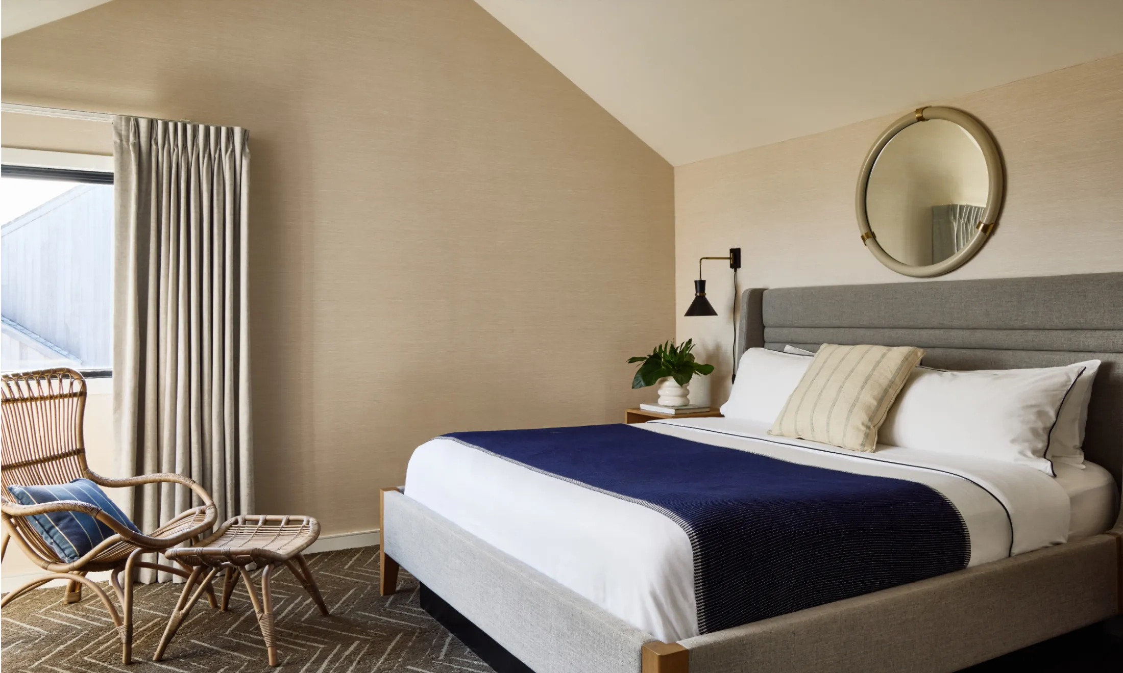 A neatly made bed with white and blue linens, a gray upholstered headboard, and a round mirror above it, with a chair and small table beside a window.