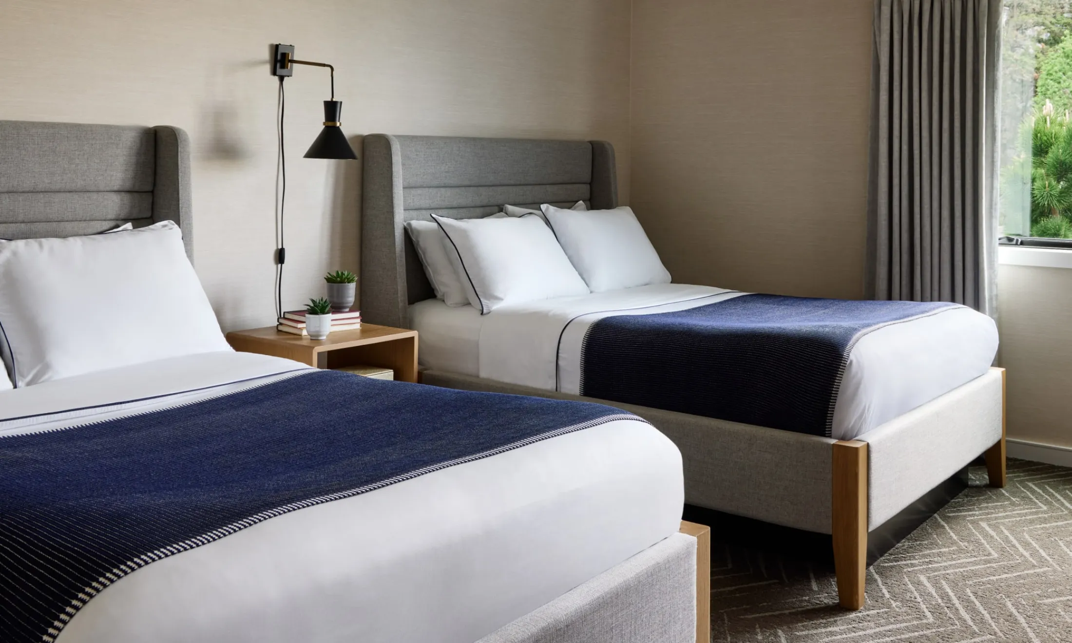 A hotel room with two twin beds, a nightstand with a lamp and a potted plant, and a window with a view of greenery outside.