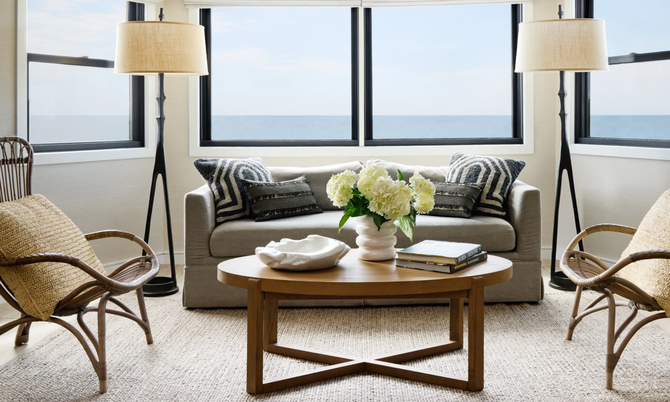 A living room with a gray sofa, two wicker armchairs, a wooden coffee table, and two floor lamps, with a large window showing an ocean view.