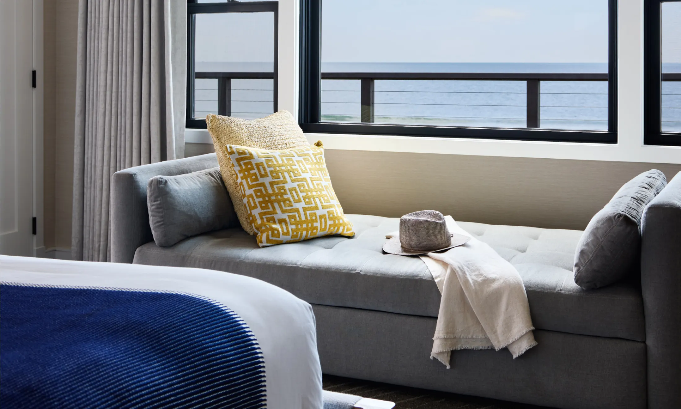 A living area with a gray couch, yellow and white pillows, and an ocean view through large windows.