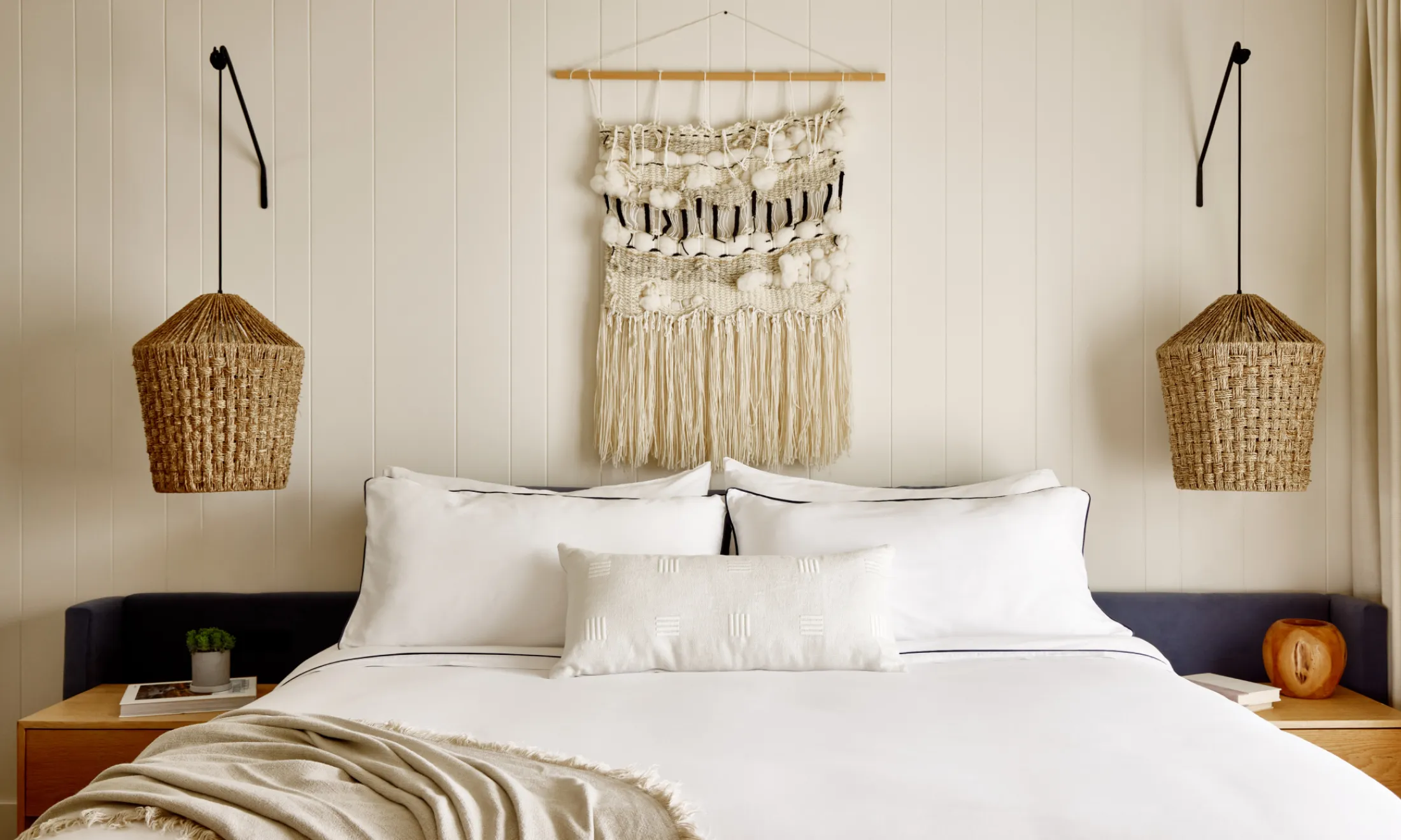 A neatly made bed with white linens and pillows, flanked by nightstands and hanging lamps, with a wall decoration above the bed.