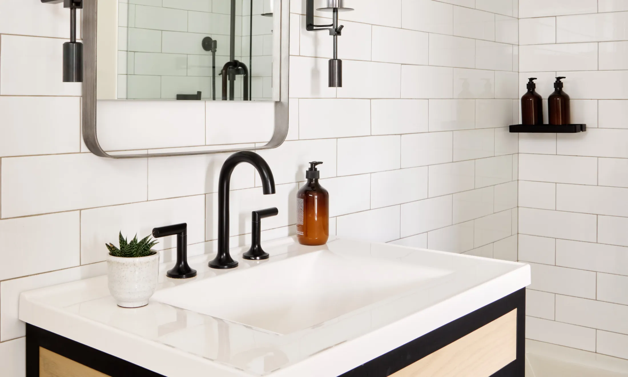 A clean bathroom sink area with white tile, a black faucet, and a small plant.