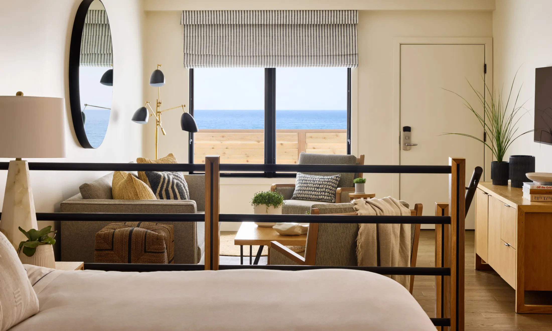 A wide view of a hotel room with a partial ocean view, featuring a bed, sofa, armchair, coffee table, and TV.