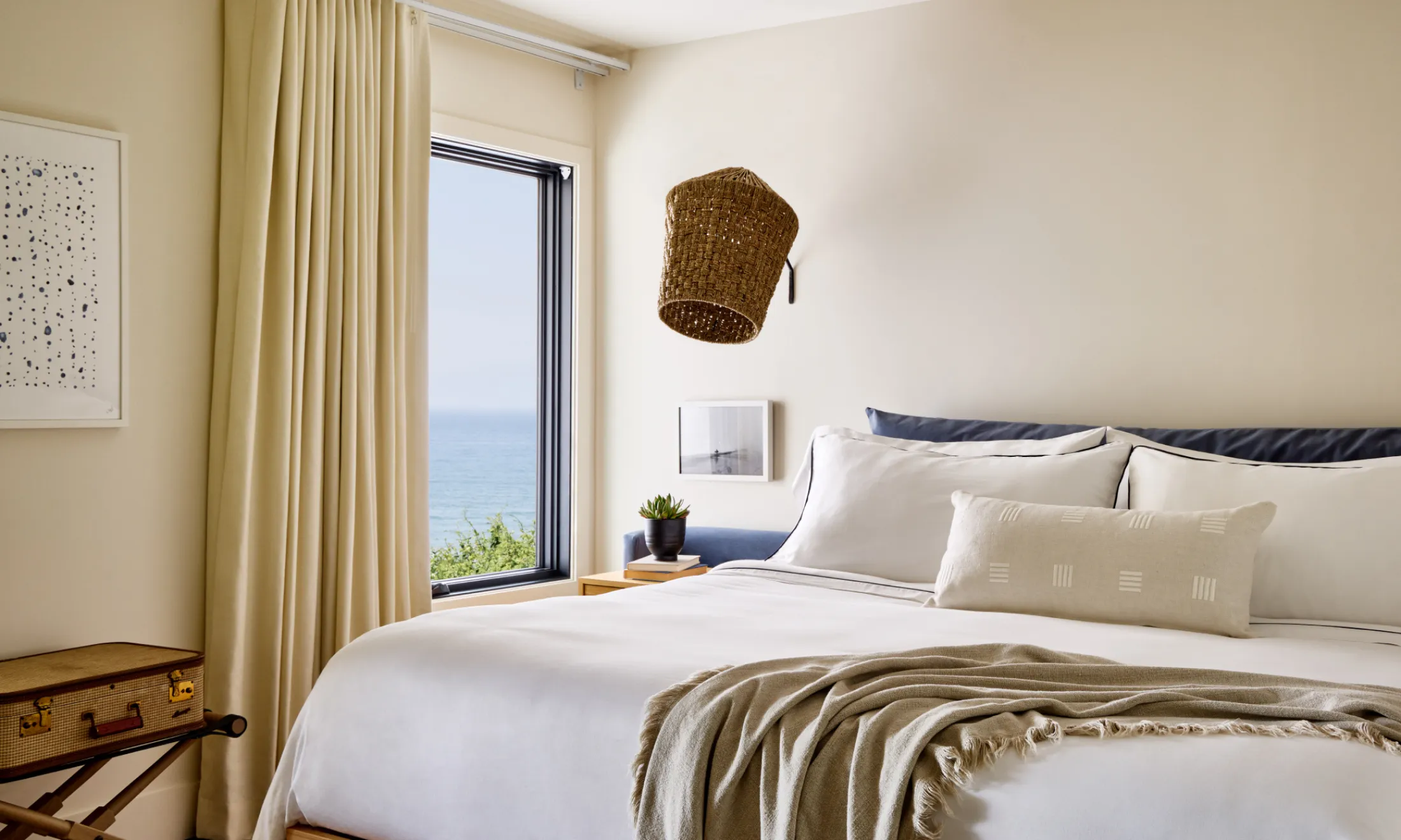 A neatly made bed with white linens and a blue headboard is positioned in front of a window with a view of the ocean.