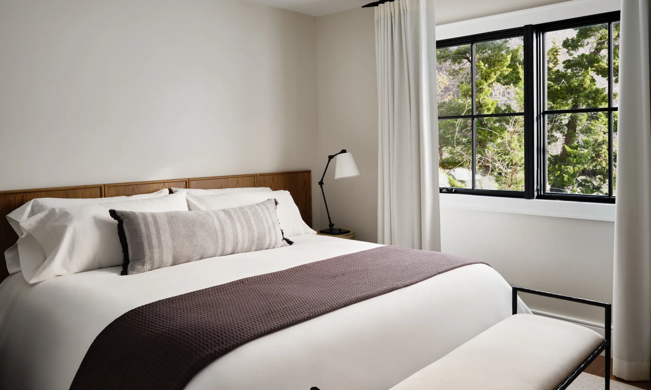A neatly made bed with white sheets and pillows, a brown throw blanket, and a wooden headboard stands in front of a window with a view of trees.