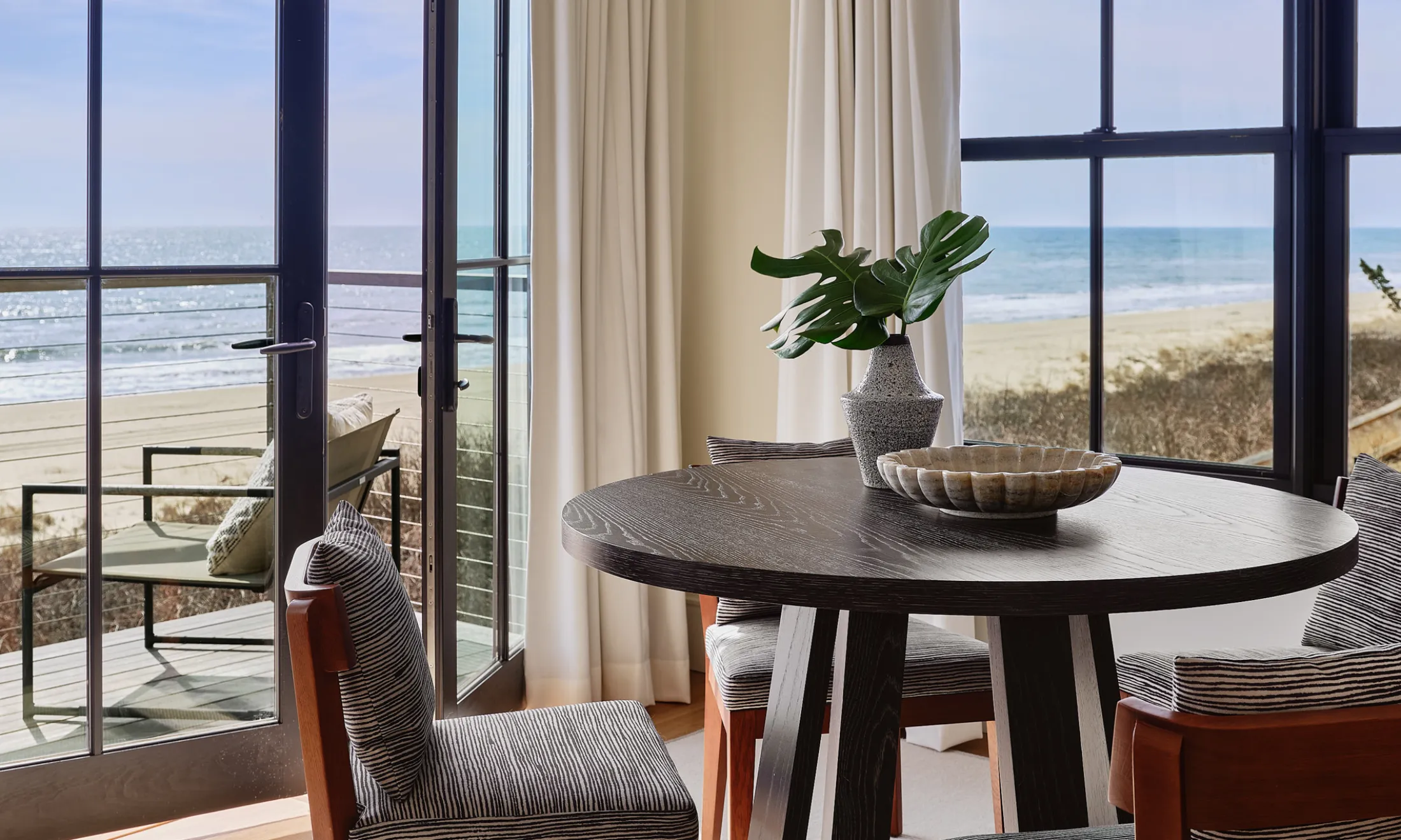 A dining area with a round table and chairs, overlooking the beach through large windows.