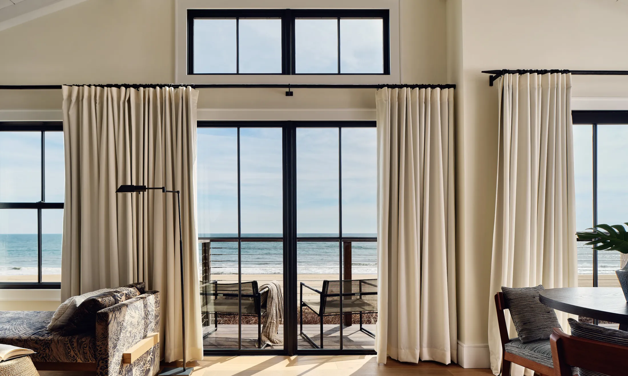 A wide room with a beach view, showing a sofa, dining table, and curtains.
