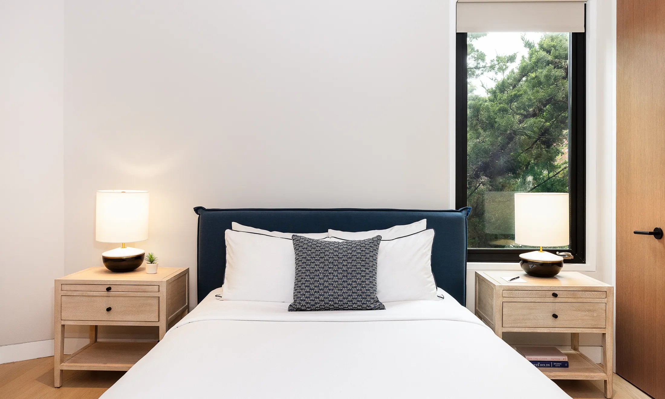 A neatly made bed with a dark blue headboard, white linens, and a black-and-white patterned pillow, flanked by two light wood nightstands with lamps, in front of a window showing trees outside.
