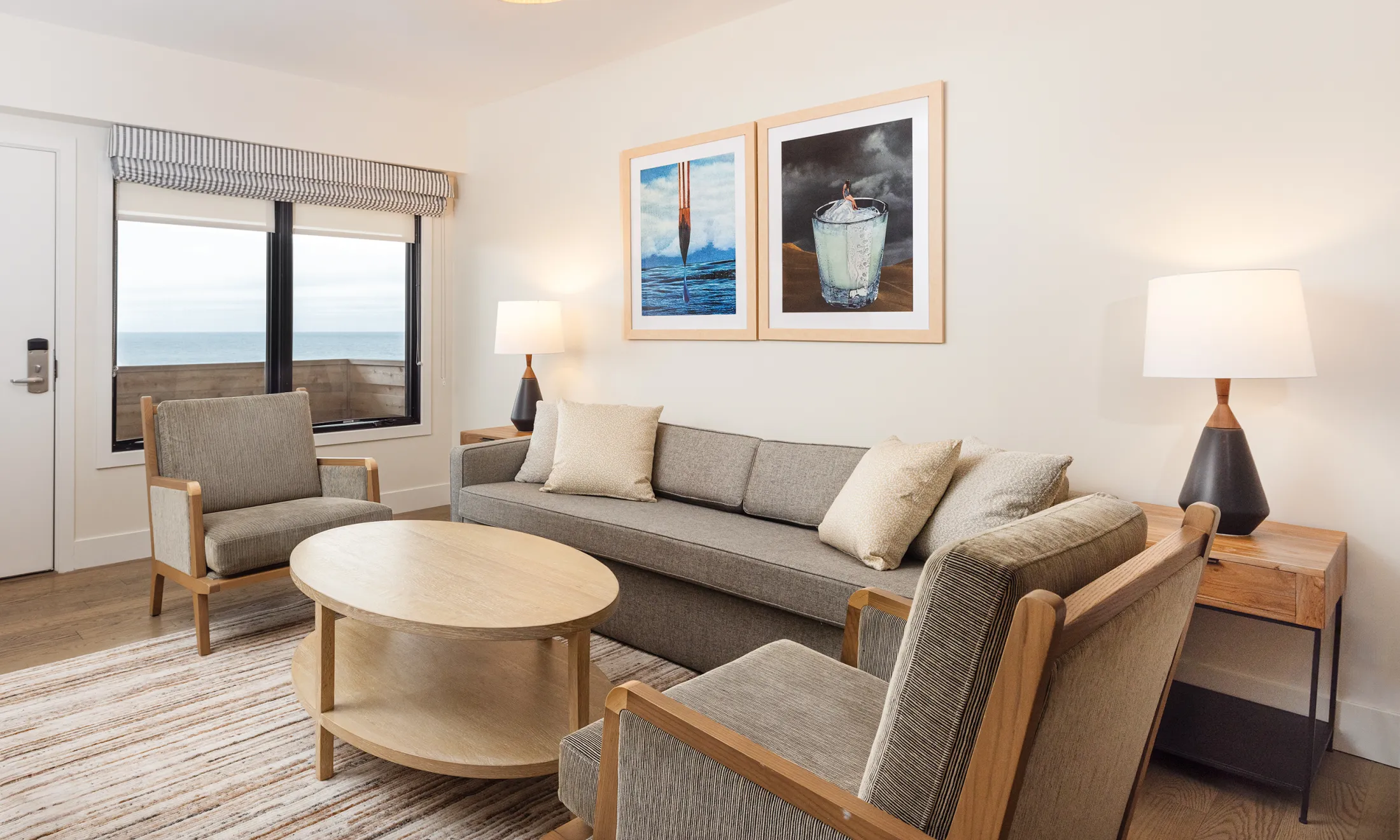 A furnished living room with a sofa, armchairs, coffee table, lamps, and artwork, overlooking the ocean through a window.