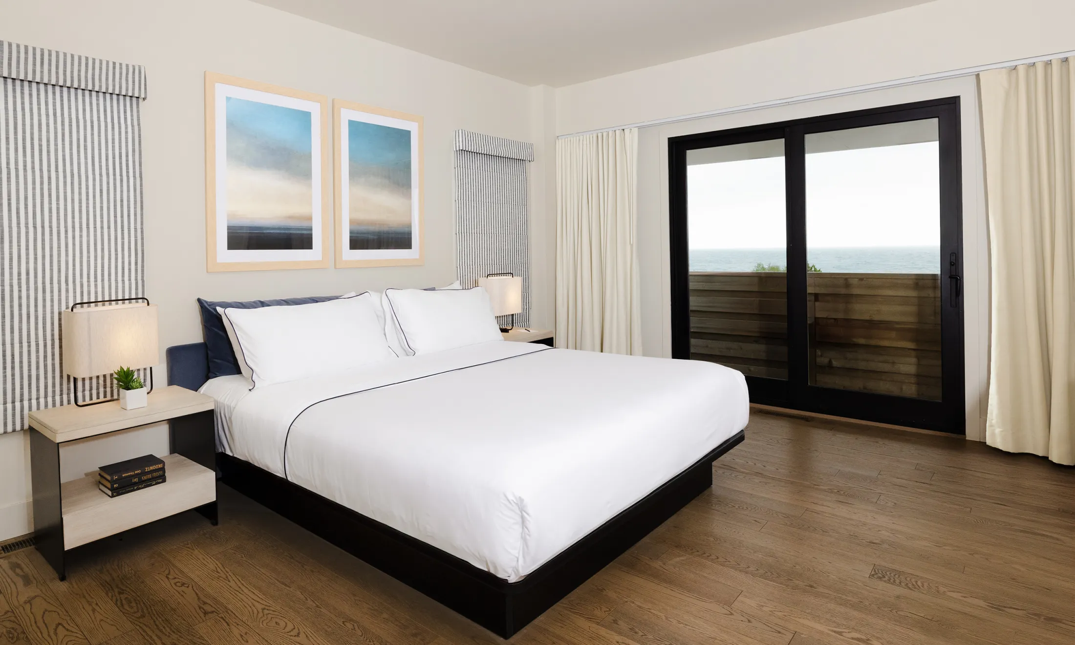 A hotel room with a double bed, nightstand, lamp, and artwork, with a sliding glass door leading to a balcony with an ocean view.