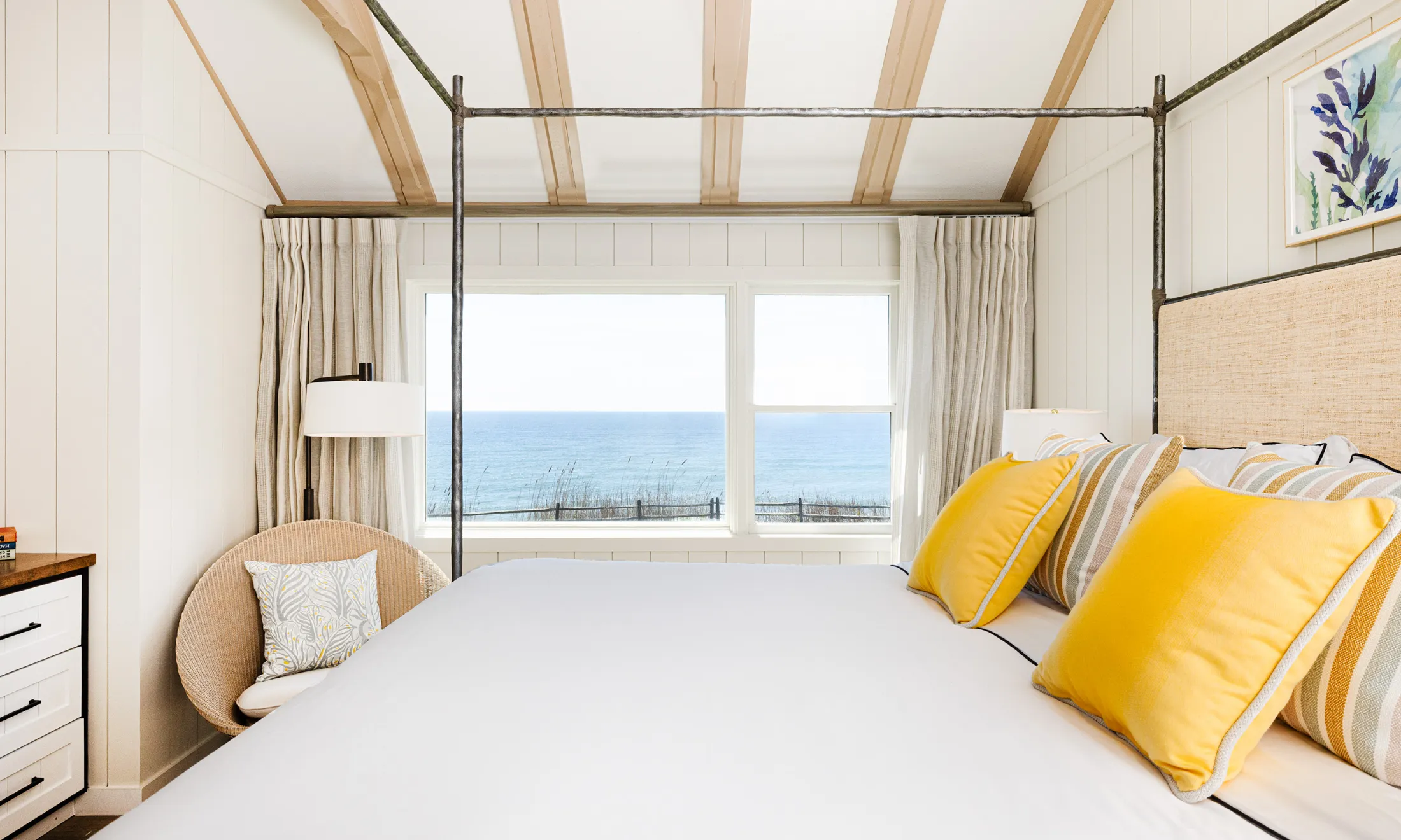 A wide view of a bright and airy bedroom with a large bed, a window with ocean views, and a comfortable reading chair.