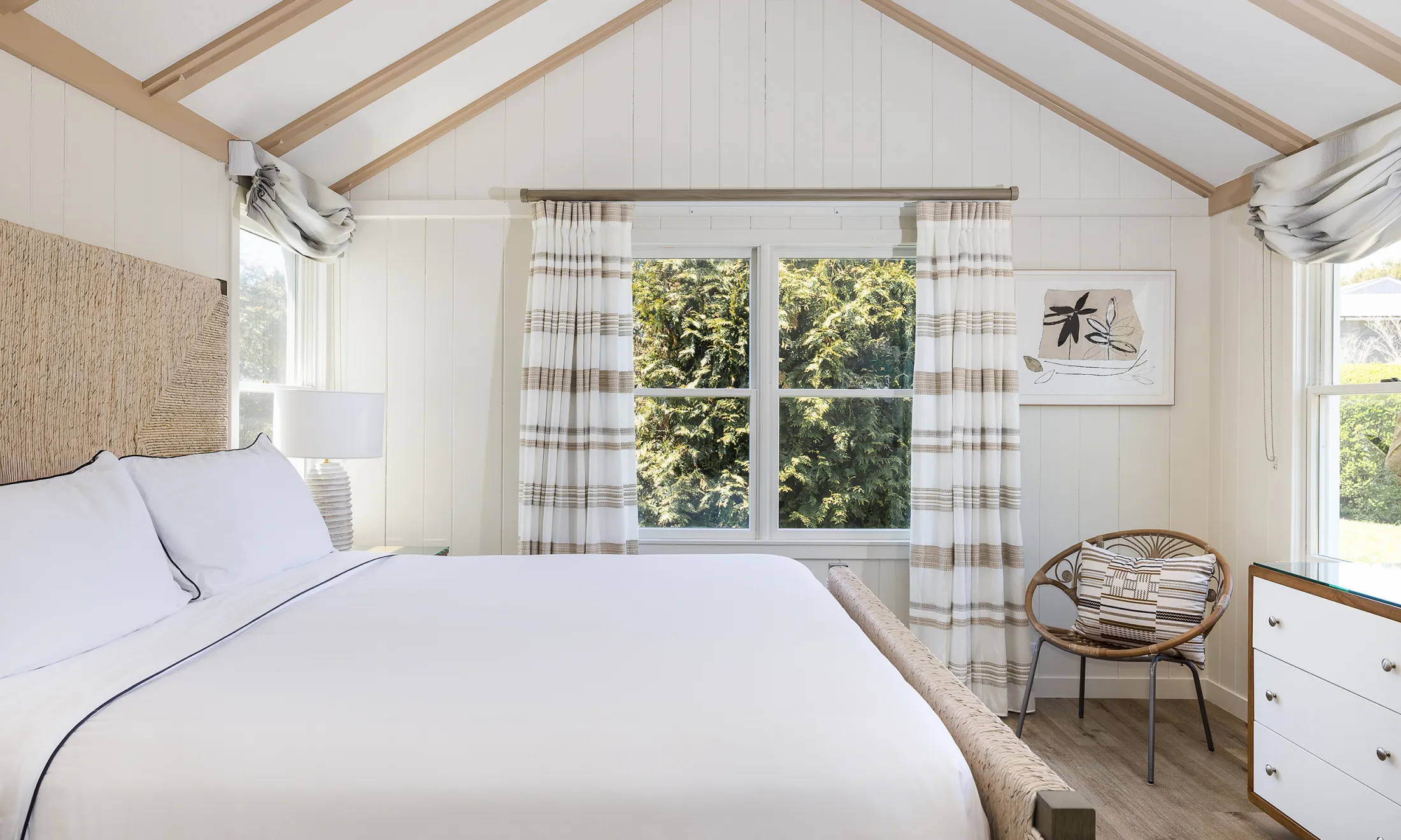 A white bedroom with a vaulted ceiling, a large bed, and a window with a view of trees.