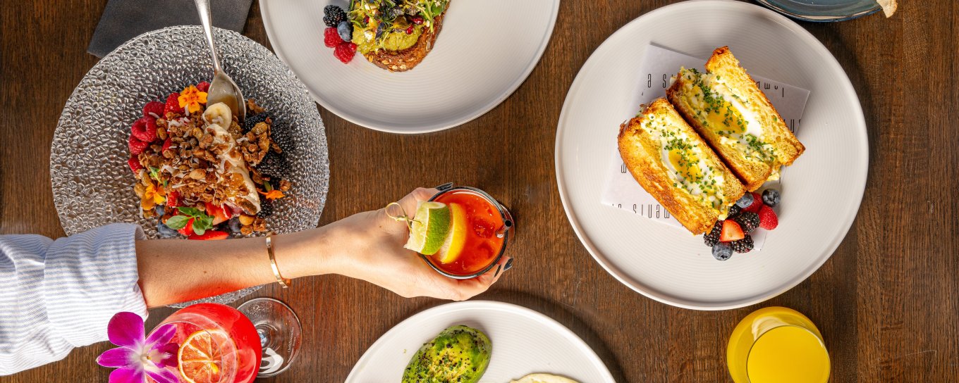 A table with various brunch dishes, including avocado toast, sandwich, and desserts, with a hand reaching for a drink with a lime garnish.