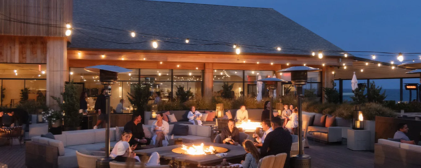 Outdoor restaurant patio at dusk with people gathered around glowing fire pits, surrounded by string lights and modern seating.