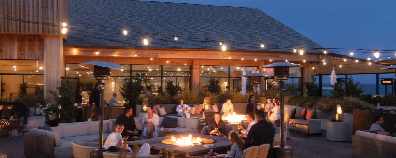 Outdoor restaurant patio at dusk with people gathered around glowing fire pits, surrounded by string lights and modern seating.