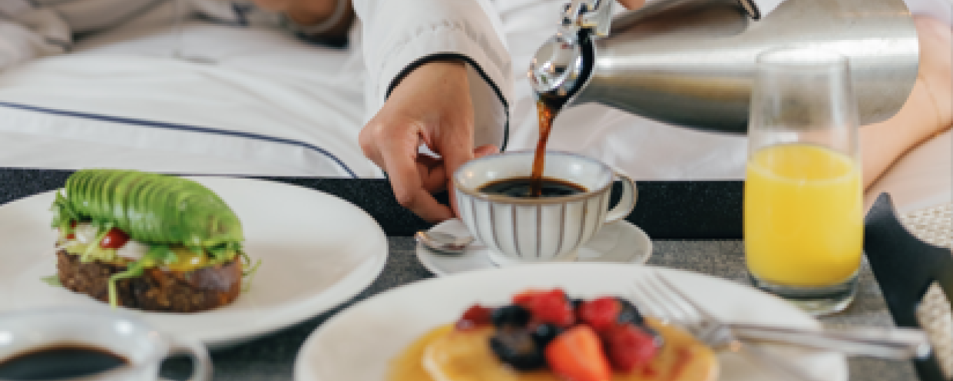 Breakfast in bed with pancakes, berries, avocado toast, coffee being poured, and orange juice.