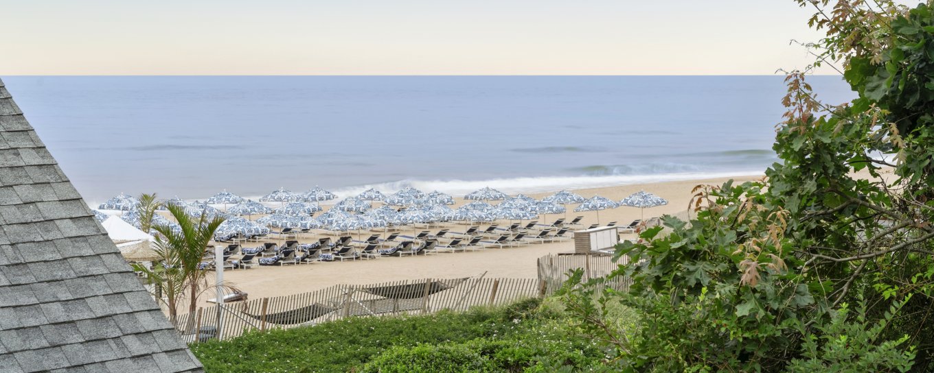 Ocean view with beach and lounge chairs seen from a lush garden and house roof. Blue sky and calm sea in the background.
