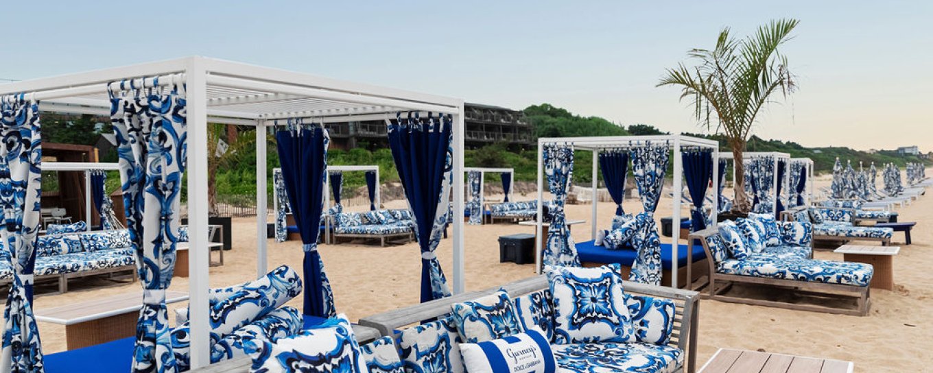 Beachside cabanas with blue patterned cushions and curtains, set on sandy shore with a clear sky and a solitary palm tree.