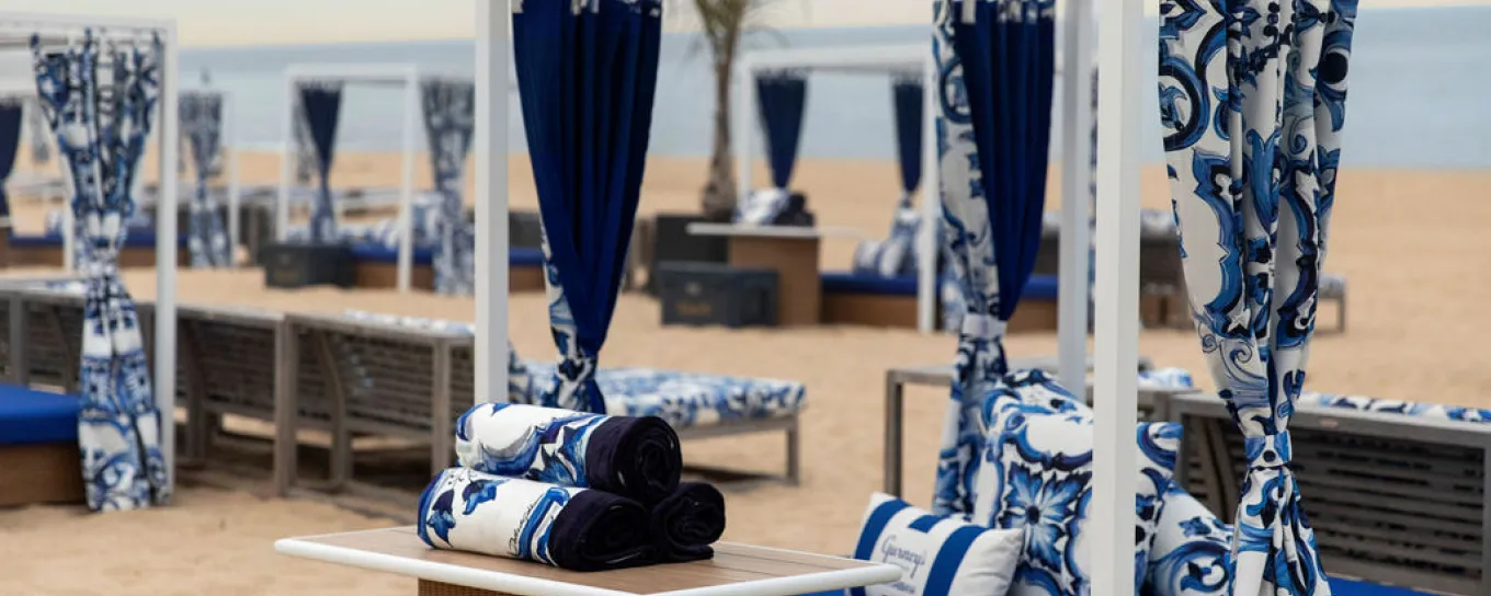 Beach cabanas with blue and white patterned curtains and cushions on sandy beach, ocean in the background.