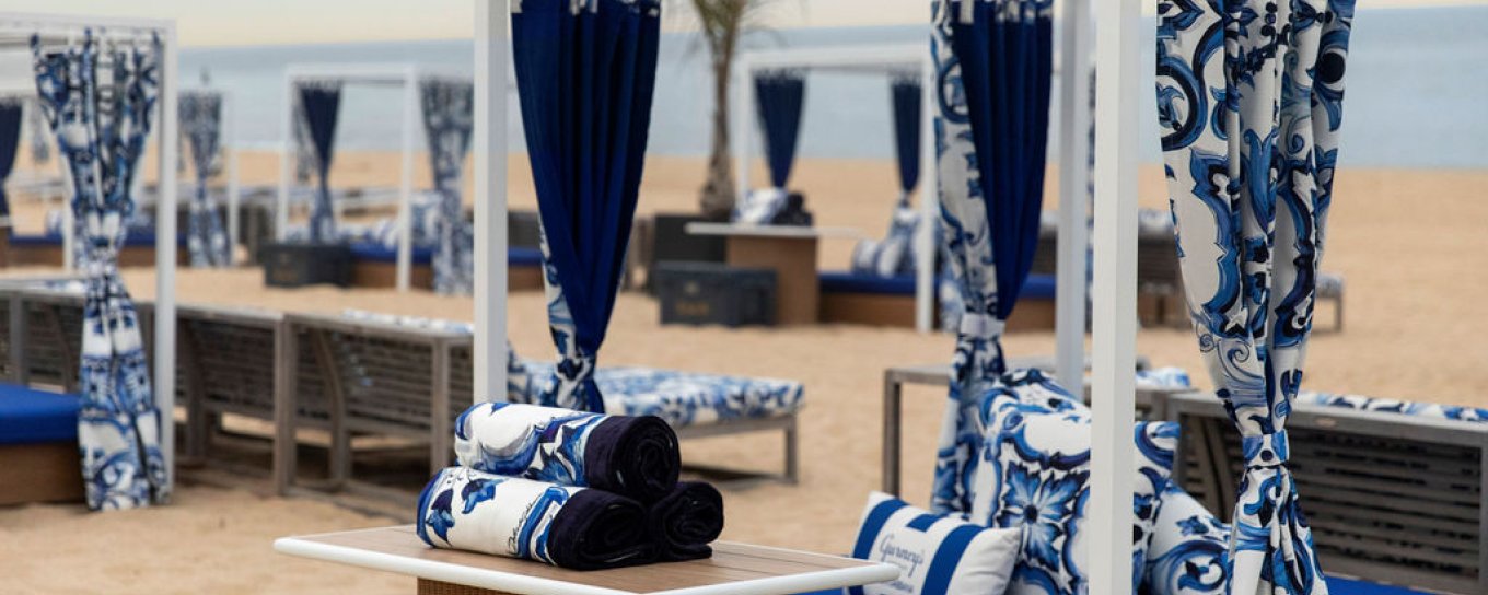 Beach cabanas with blue and white patterned curtains and cushions on sandy beach, ocean in the background.