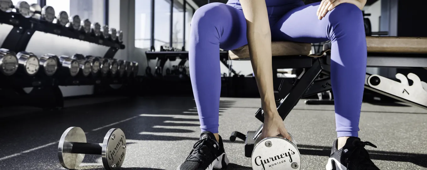 Woman in gym attire seated on a bench, holding a medicine ball labeled "Garden of Life," with dumbbells on the floor nearby.
