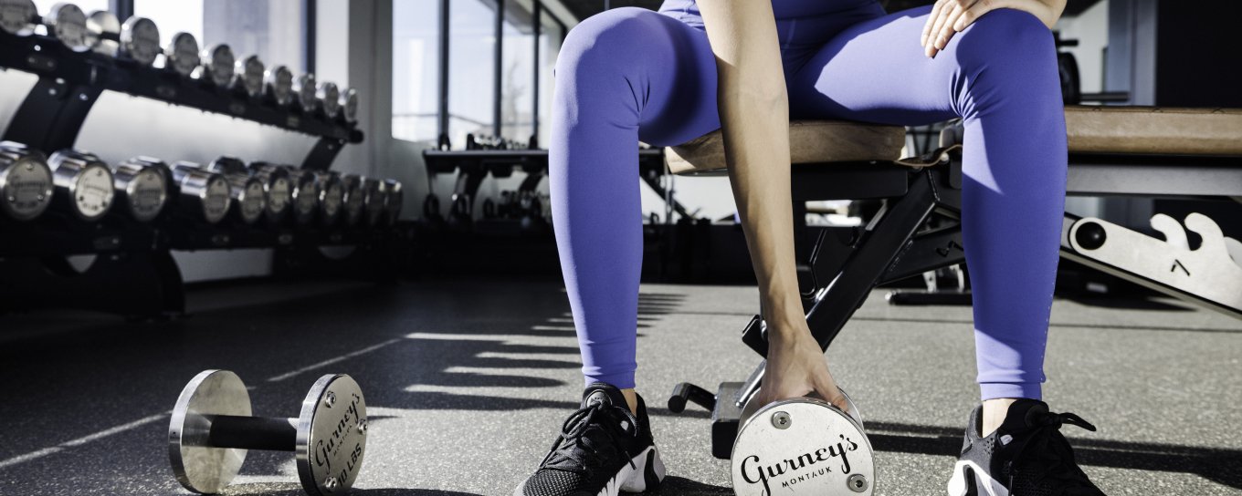 Woman in gym attire seated on a bench, holding a medicine ball labeled "Garden of Life," with dumbbells on the floor nearby.