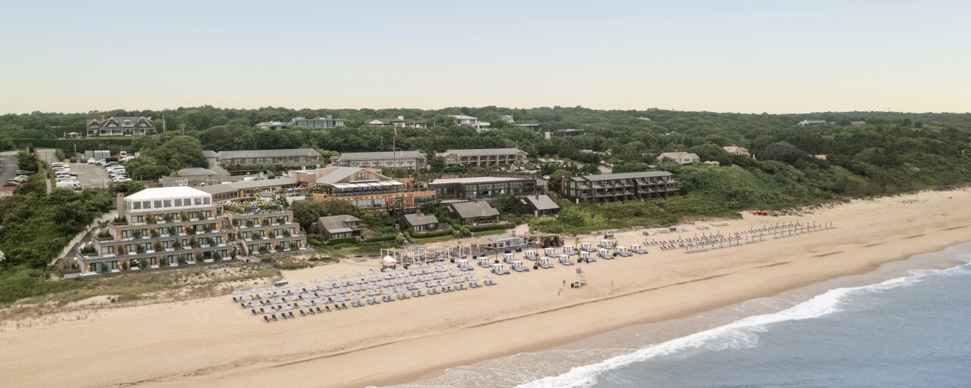 Aerial view of a beach with rows of umbrellas, buildings set back among green trees, and gentle waves on the sandy shore under a clear sky.