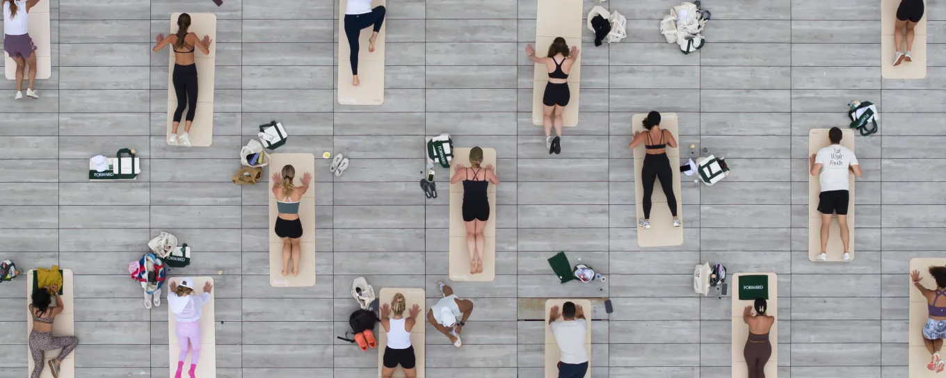 People lying on yoga mats in neat rows on a gray deck, viewed from above, some with bags beside them.
