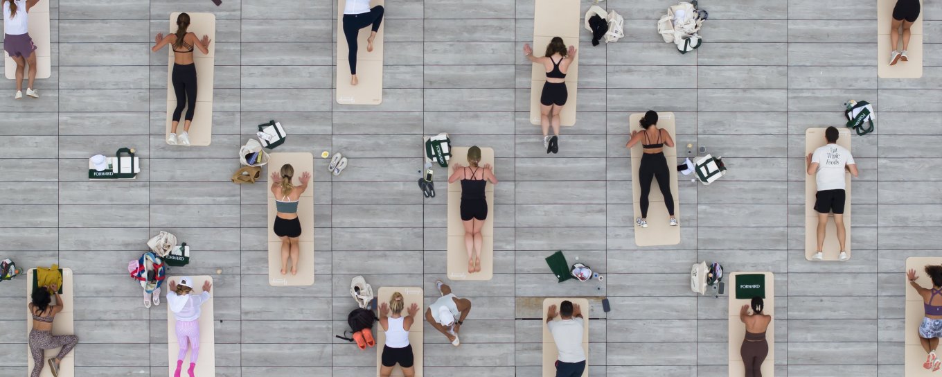 People lying on yoga mats in neat rows on a gray deck, viewed from above, some with bags beside them.