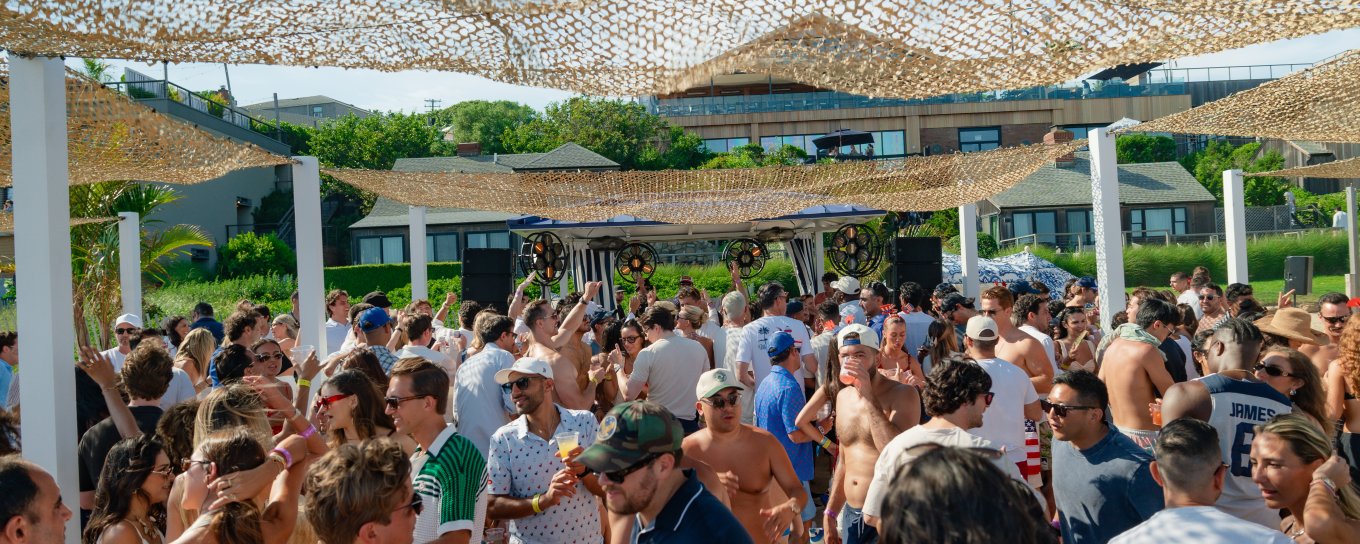 Crowded outdoor party with people mingling under woven canopies. Sunny day with greenery and buildings in the background.