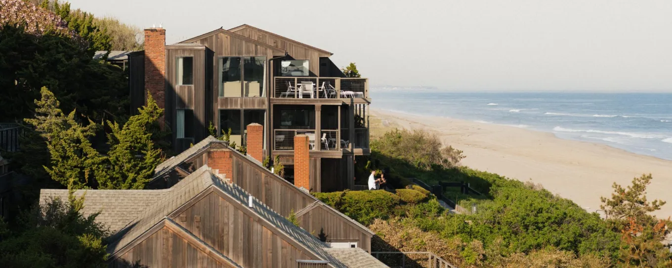 Wooden beach houses nestled among trees with a sandy beach and ocean stretching into the distance under a clear blue sky.