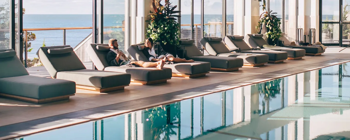 Indoor pool area with lounge chairs, large windows overlooking the ocean, and potted plants. Sunlight reflects on the water.