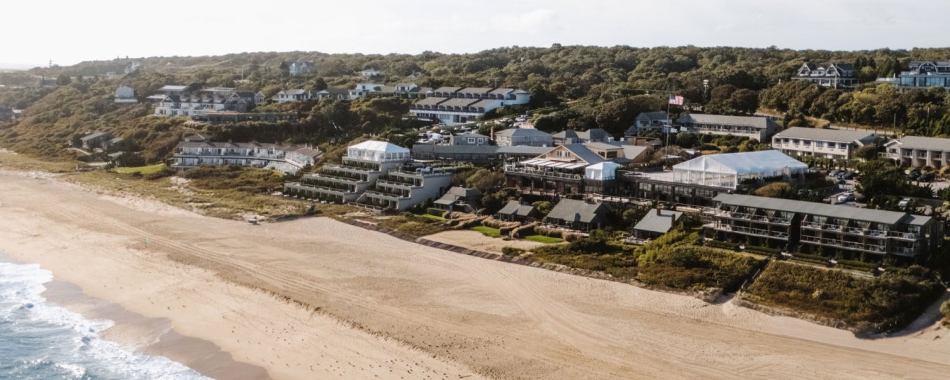 Aerial view of a sandy beach with waves, backed by a coastal town with green trees and buildings in the background.