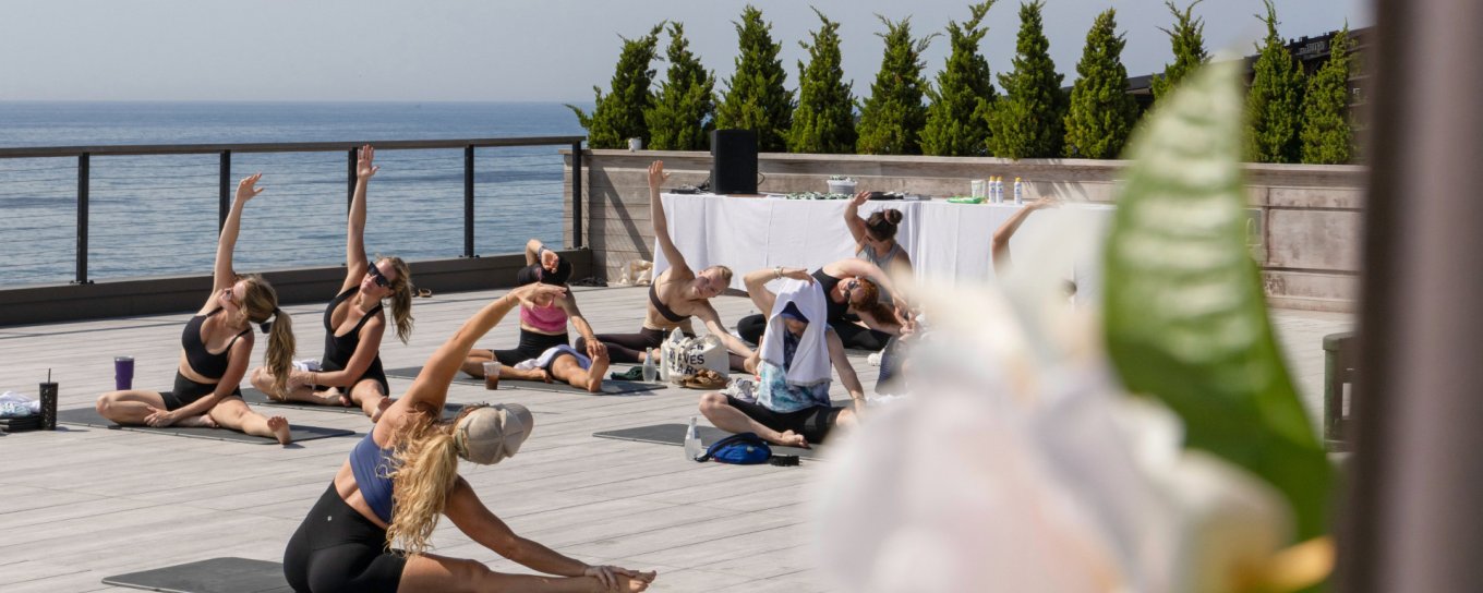 People practicing yoga outdoors on a wooden deck, overlooking the ocean. Greenery and blue sky in the background.