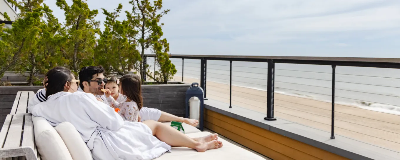Family relaxing on a terrace overlooking the beach, wearing white robes. The ocean and blue sky are in the background.