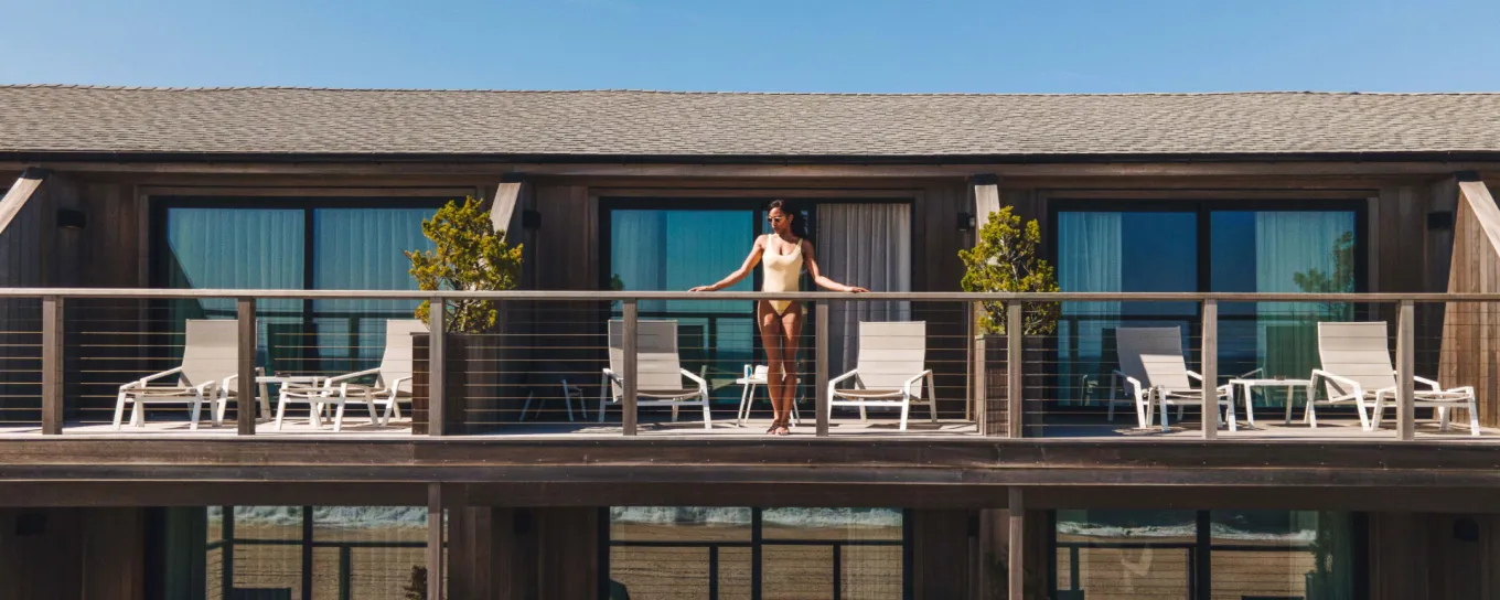 A person stands on a wooden balcony surrounded by white chairs and potted plants, with blue sky above, attached to a two-story building.