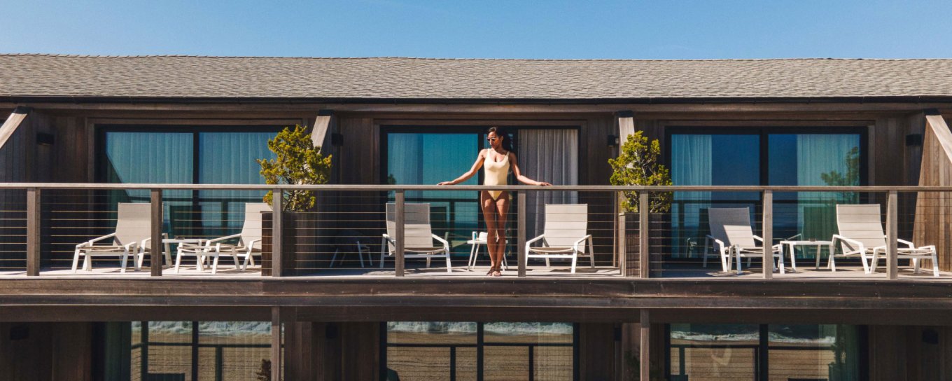 A person stands on a wooden balcony surrounded by white chairs and potted plants, with blue sky above, attached to a two-story building.