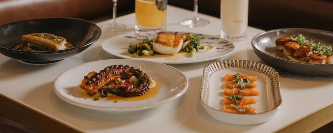 Assorted gourmet dishes on a restaurant table, including seafood, vegetables, and drinks in the background.