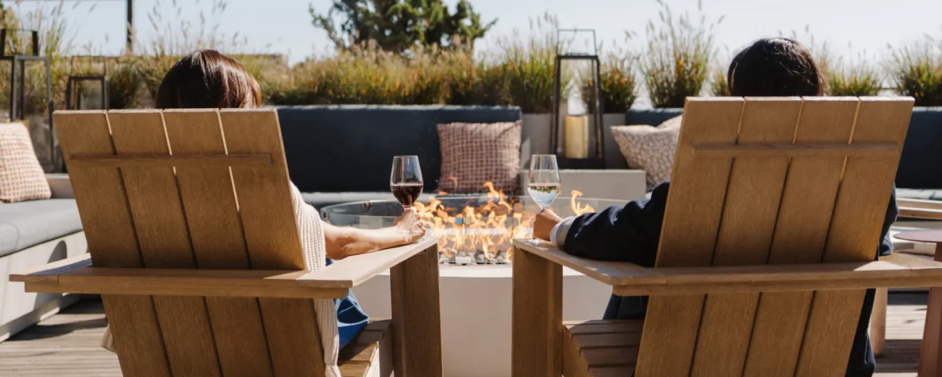 Two people relax in wooden chairs by a fire pit, holding glasses of wine on a sunny patio.