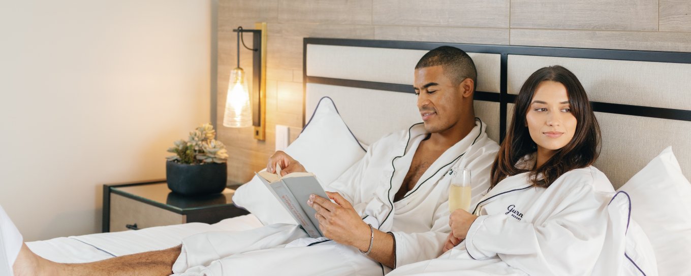 Couple in bathrobes relax in bed, enjoying breakfast on a tray with juice and fruit, in a bright, modern bedroom.