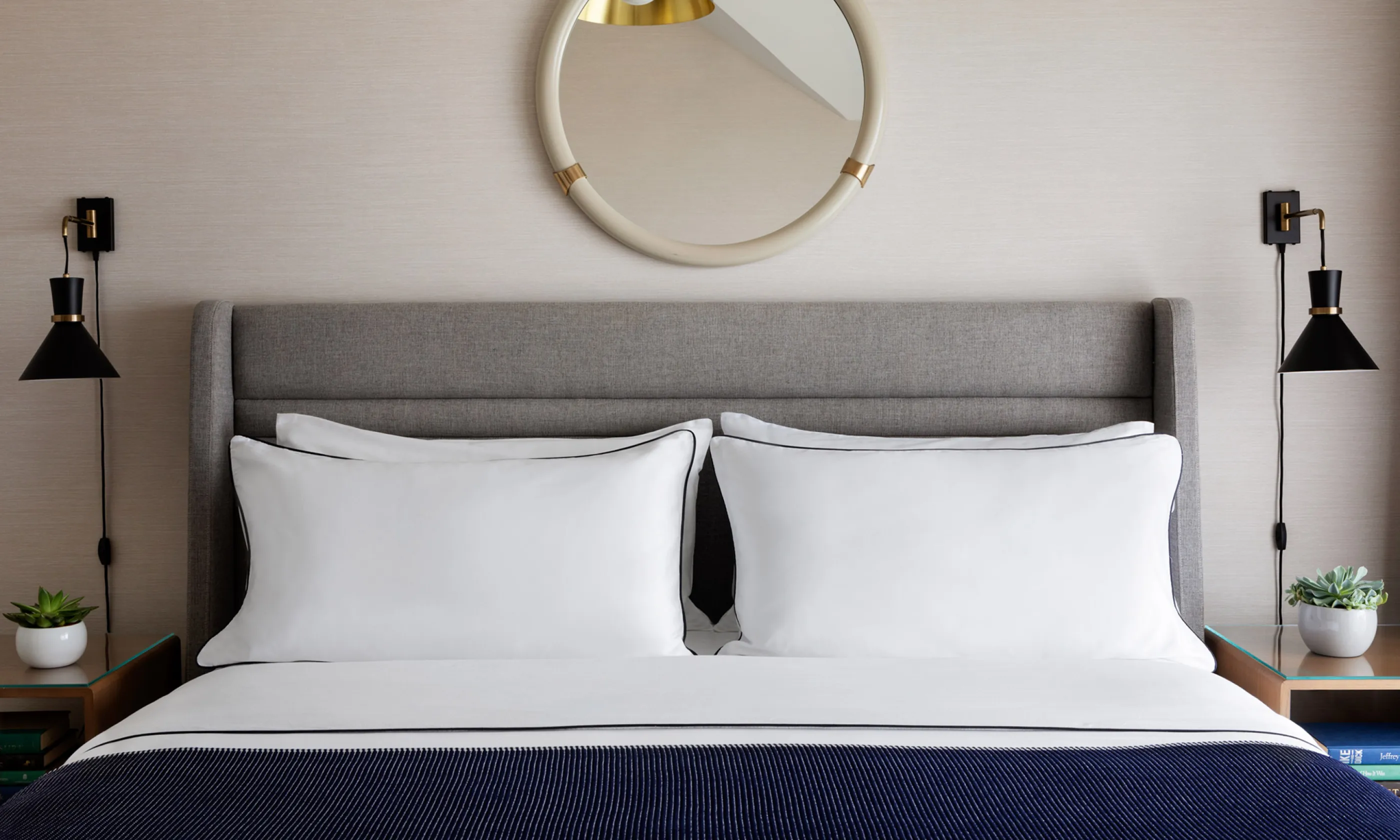 Stylish bedroom with a grey upholstered headboard, crisp white pillows, and a navy blanket. Black wall sconces and potted plants on wooden nightstands.