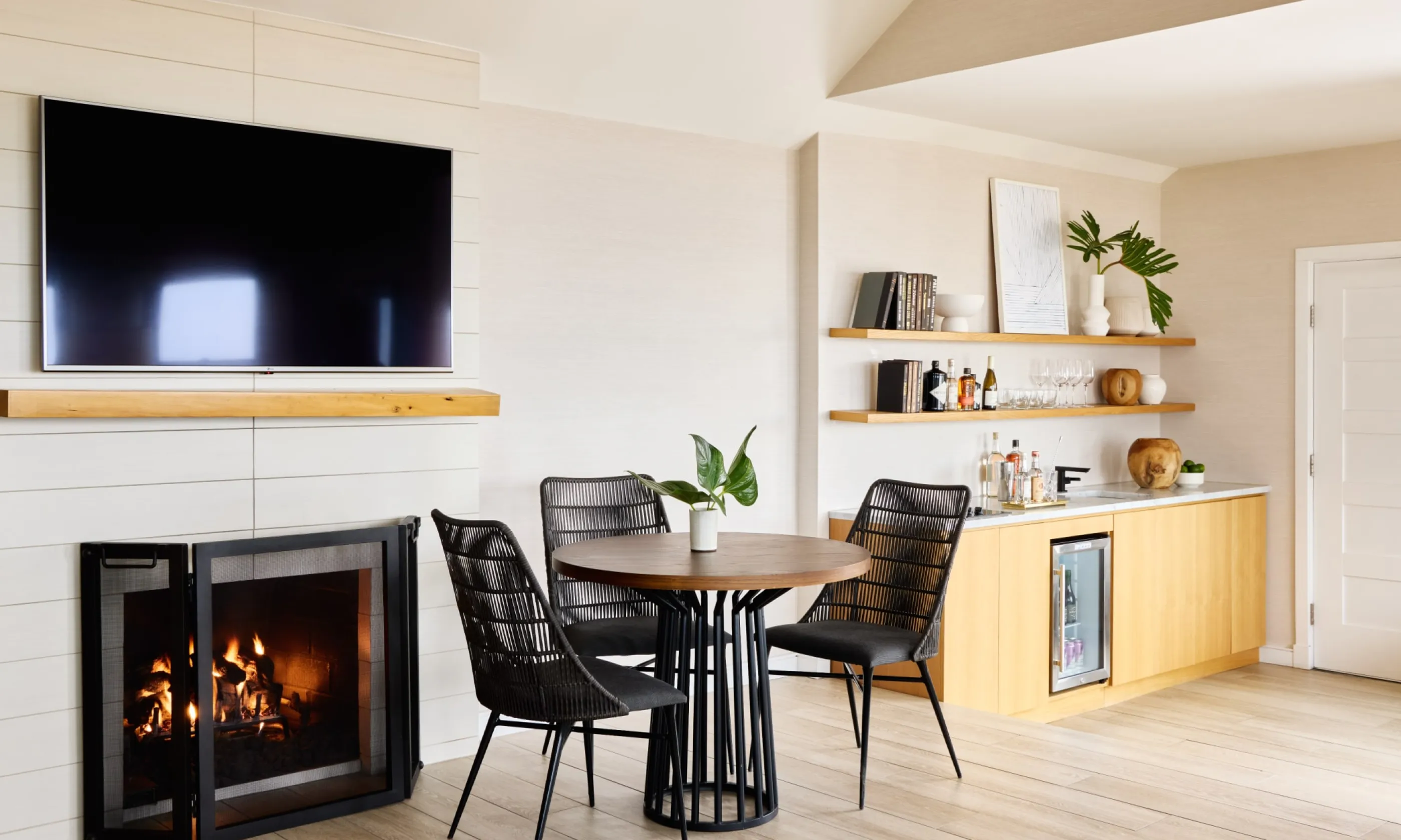 Cozy living area with a fireplace, TV, round table, and chairs. Light wood flooring and built-in shelves with decor on the right.