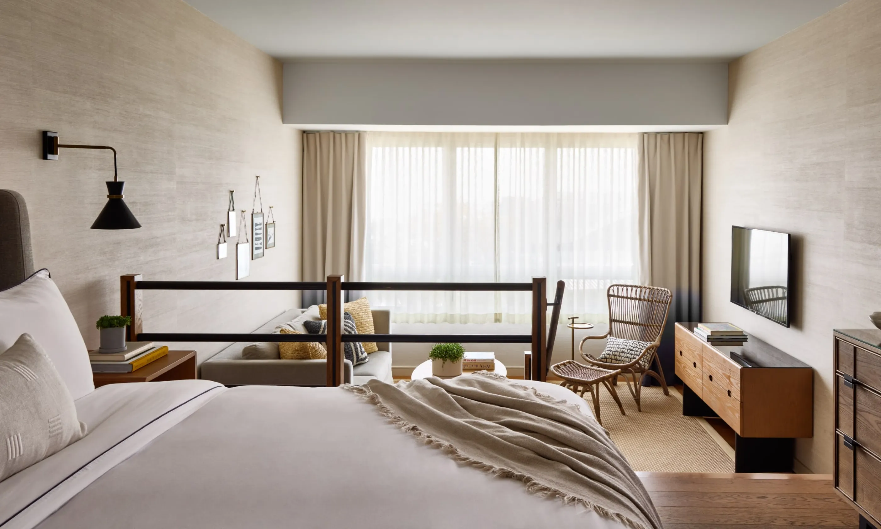 Modern hotel room with a neatly made bed, wall-mounted lamp, wood sideboard, armchair, and large window with light curtains.