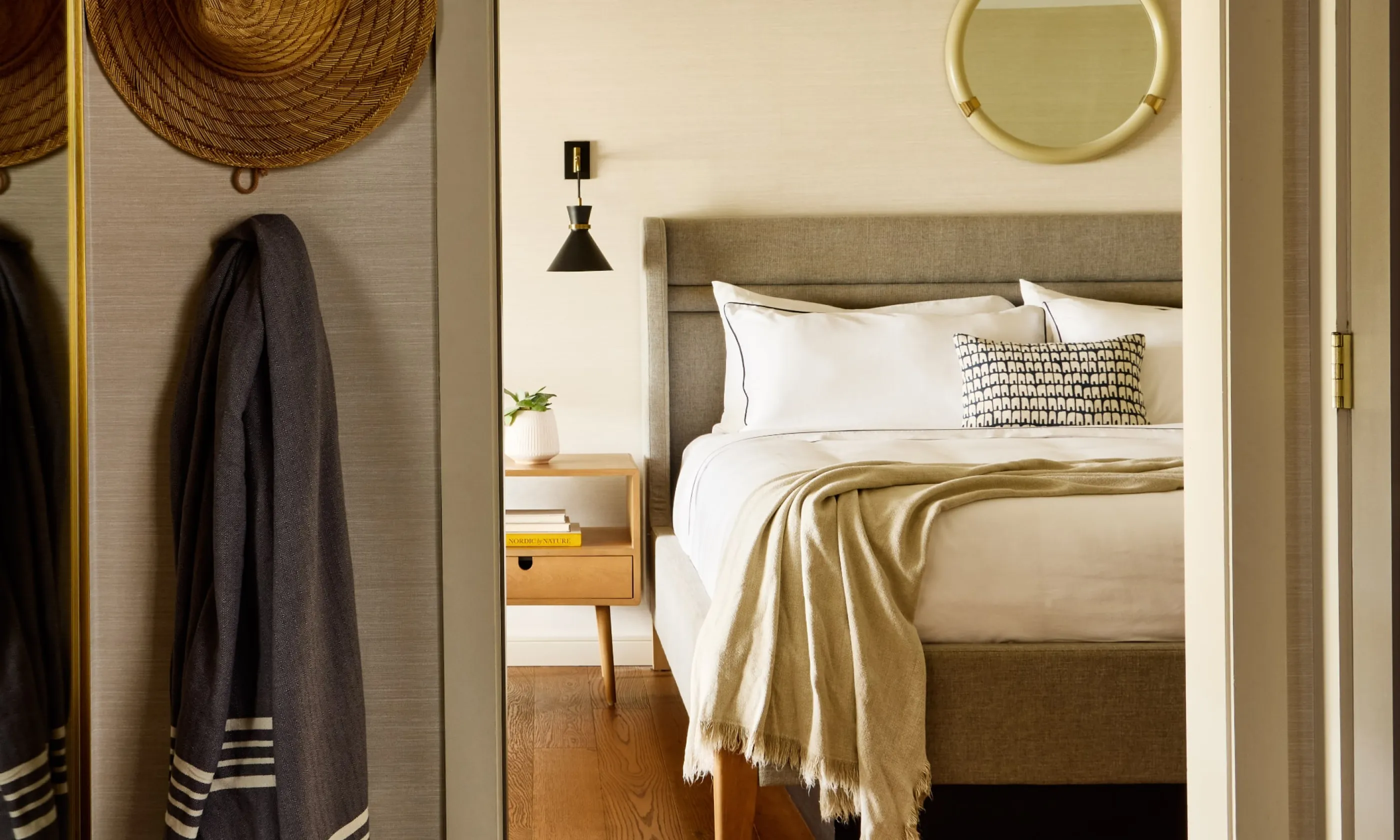 Cozy bedroom with a neatly made bed, throw blanket, and decorative pillows. A round mirror and wall lamp above the bed, with hats hanging on the wall.