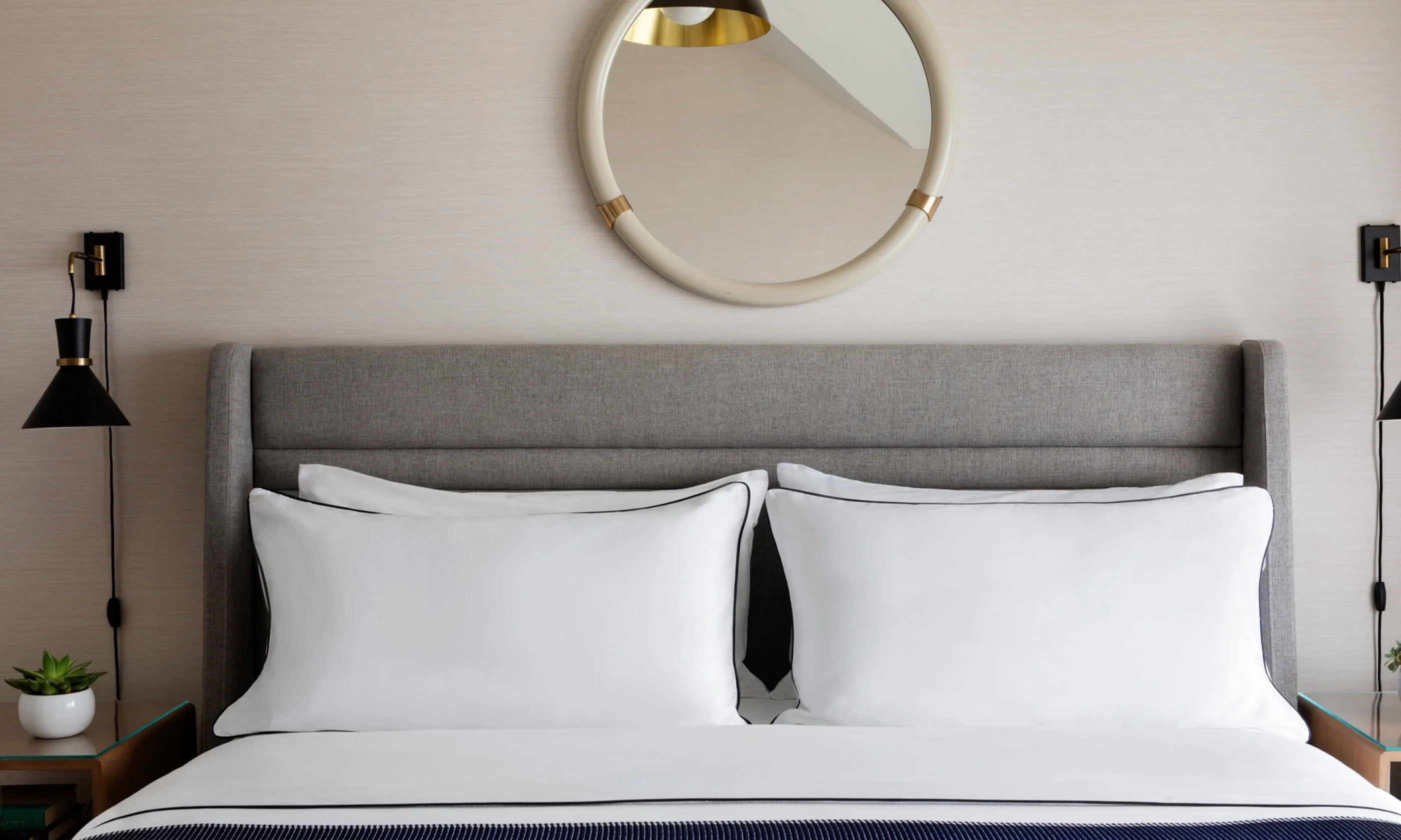 Bed with gray headboard, two large white pillows, a round mirror above, black wall lamps on each side, small plants on nightstands.